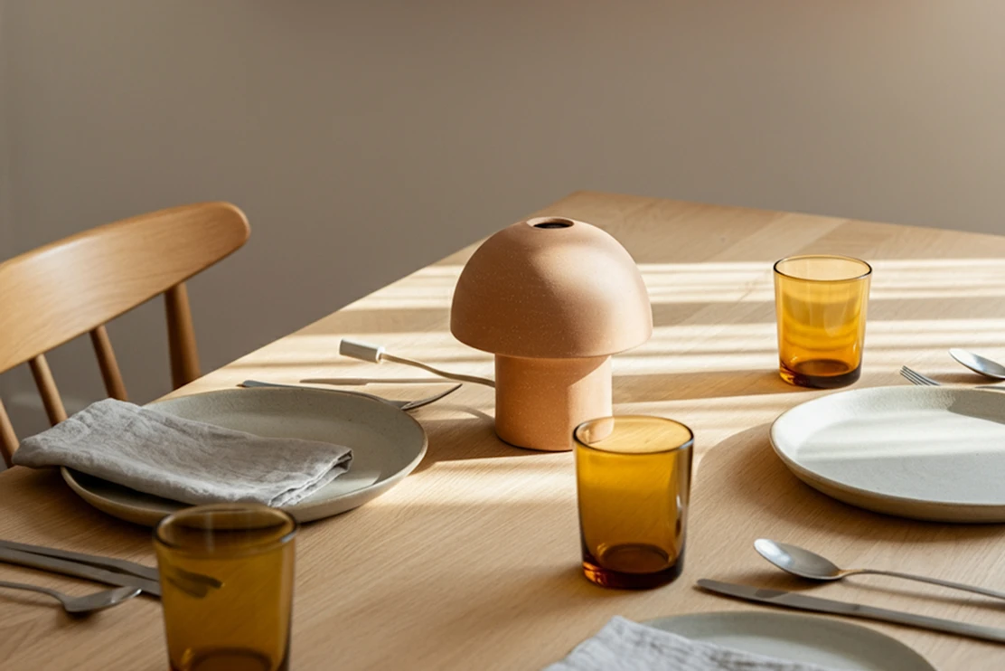 Wooden dining table set with a terracotta lamp, amber glasses and plates in warm sunlight