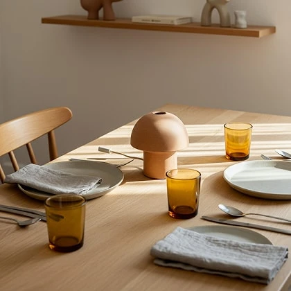 Wooden dining table set with a terracotta mushroom lamp, amber glasses and linen napkins in warm sunlight